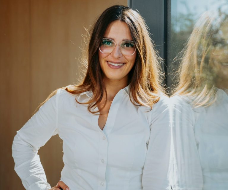 Frau mit langen Haaren in weißem Hemd lächelt, steht vor einem Fenster.